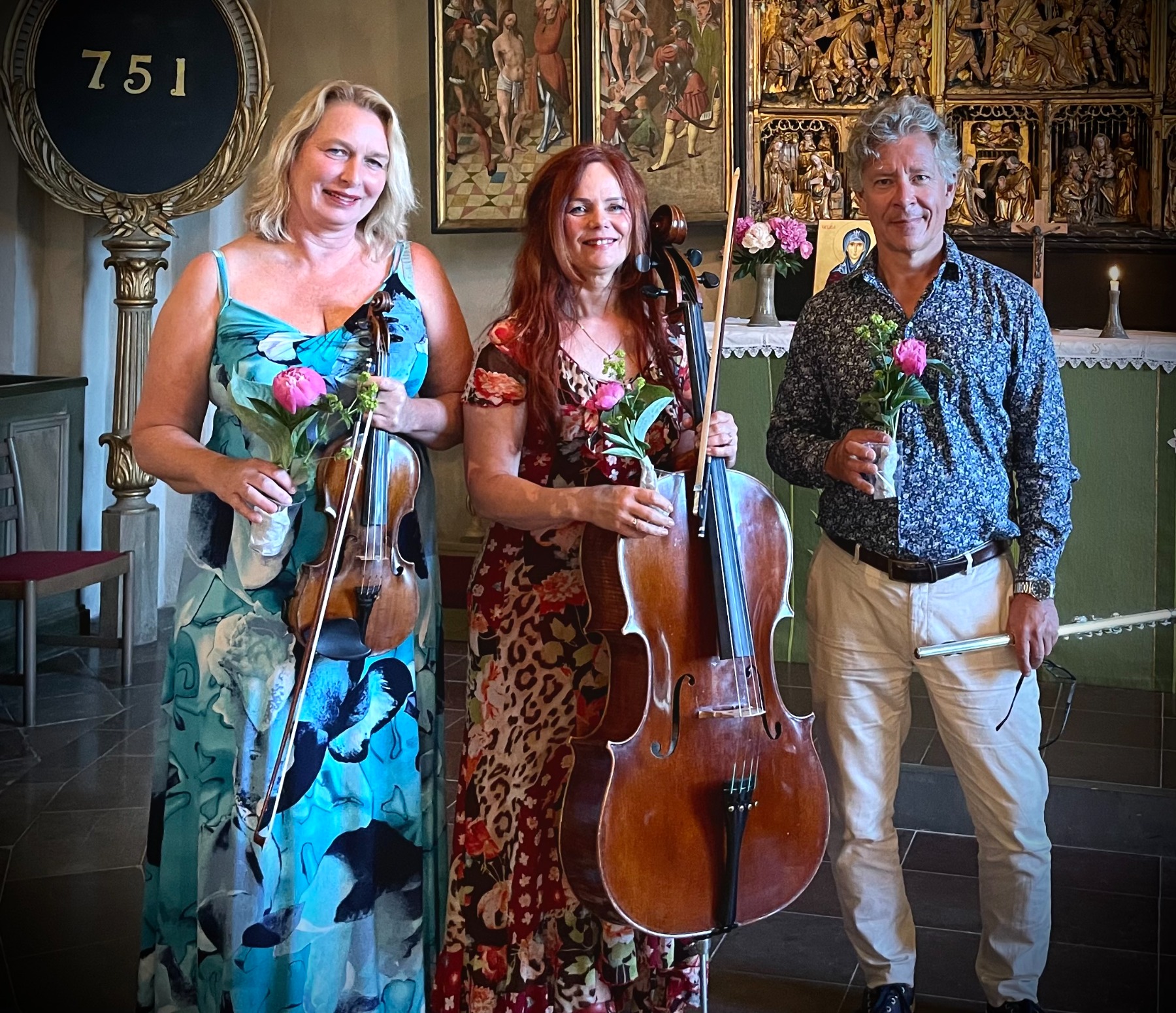 Marie Louise Sjöberg, Lena Müller och Erik Sunnerstam står leende på rad i en kyrka. De håller i sina instrument och varsin pion.