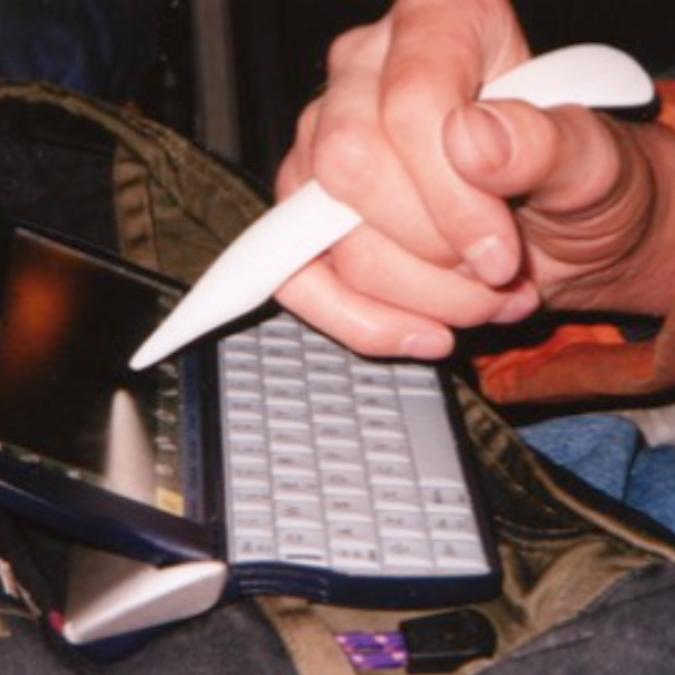 A hand holding a white stylus between the middle- and ring finger, pointed at a mini computer screen.