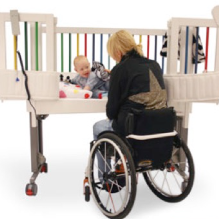 En spjälsäng där ena långsidan är försedd med dörrar som öppnats upp. En person i en rullstol sitter bredvid och leker med ett leende barn som ligger i spjälsängen.