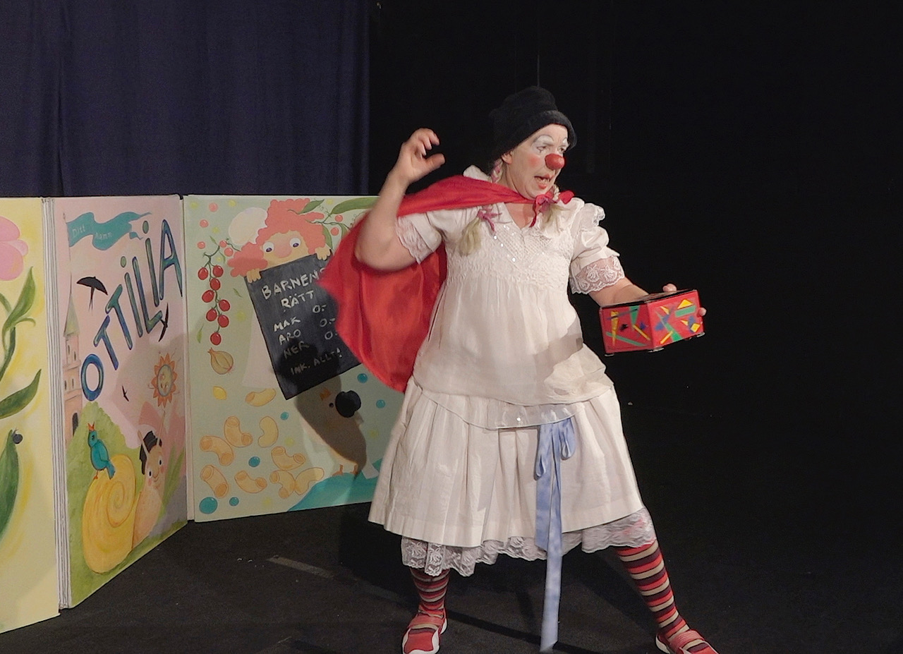 Clownen Ottilia med svart hatt, röd slängkappa, och i handen ett färgglatt skrin.